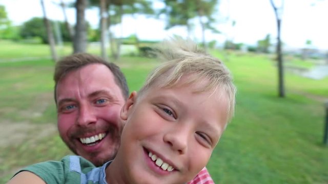 Portrait of happy caucasian family spinning around while taking selfie in green pine park. Laughing daddy and son posing for camera on summer hot day. Real time wide angle full hd video footage.