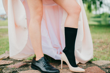 Unrecognizable woman naked legs in men sock black leather footwear. Unknown bride in expensive wedding dress and high heels creamy shoe. Marriage day. Funky, odd, strange, ridiculous girl oudoor.