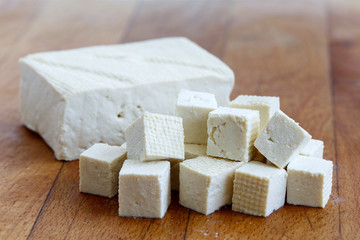 Single block of white tofu with cut tofu cubes on wooden chopping board.