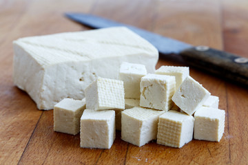 Single block of white tofu with cut tofu cubes and rustic knife on wooden chopping board.
