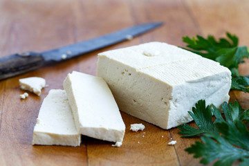 Single block of white tofu with two tofu slices, crumbs, fresh parsley and rustic knife on wooden chopping board.