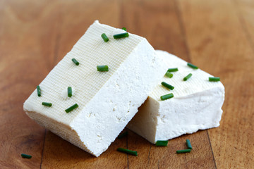 Two blocks of white tofu with fresh chives on wooden chopping board.