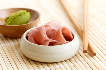 Pickled pink ginger slices in wooden bowl on bamboo mat next to chopsticks and wasabi paste in a bowl.