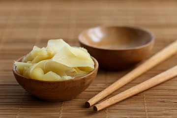 Pickled ginger slices in wooden bowl on bamboo mat next to chopsticks and empty bowl.