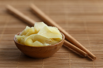 Pickled ginger slices in wooden bowl on bamboo mat next to chopsticks.