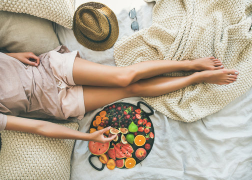 Summer healthy raw vegan clean eating breakfast in bed concept. Young girl wearing pastel colored home clothes taking fruit from tray full of fresh seasonal fruit. Top view, horizontal composition