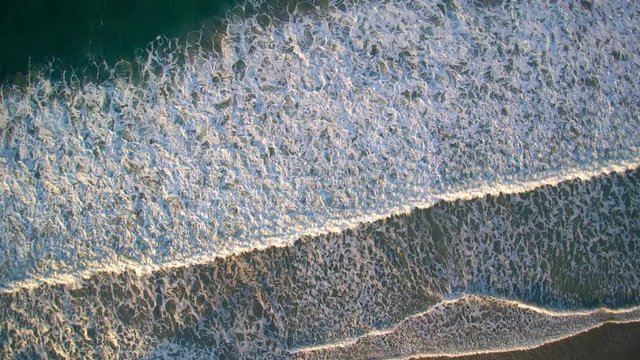 Aerial view of the seashore at golden hour - the waves crash and let foam patterns under a golden light in a sunny evening - beautiful scenery of a seascape