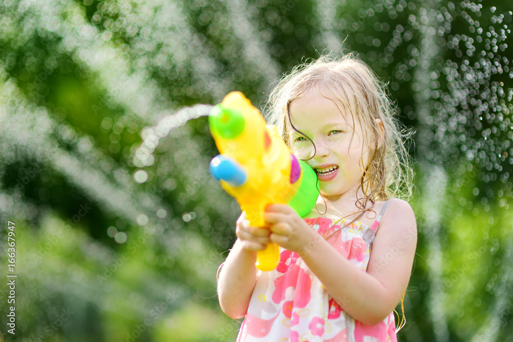 Sticker Adorable little girl playing with water gun on hot summer day. Cute child having fun with water outdoors.