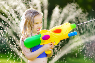 Adorable little girl playing with water gun on hot summer day. Cute child having fun with water outdoors. © MNStudio