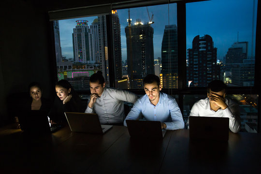 business team working late at night with lights off and computers laptop screen light on. Business people working hard concept. 20-30 year old.
