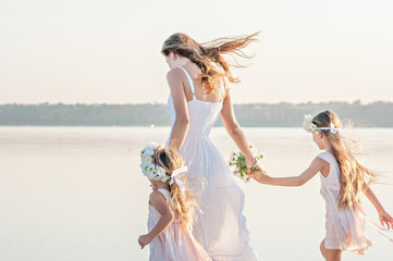 Happy Mom with two beautiful little girls walking in white dresses on the lake