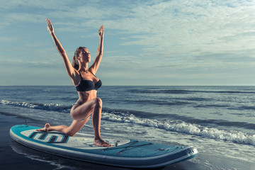 Beautiful sporty woman practicing SUP yoga at sunrise.