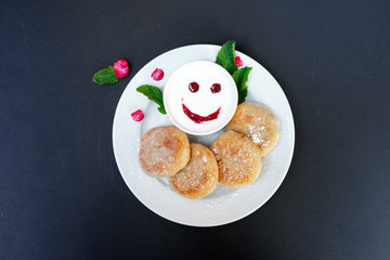 Pancakes on a plate with sour cream on a black background. Cheerful breakfast with a smile.