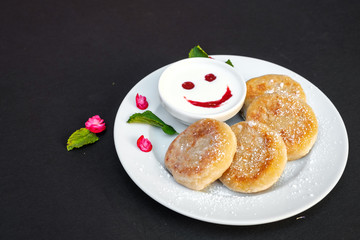 Pancakes on a plate with sour cream on a black background. Cheerful breakfast with a smile.