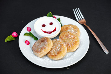Pancakes on a plate with sour cream on a black background. Cheerful breakfast with a smile.