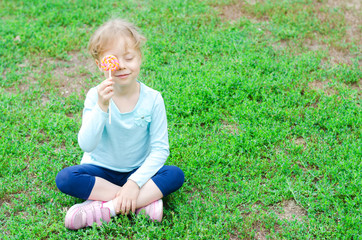 Fototapeta premium Happy little girl with a lollipop sitting on a green glade, free space for your text.