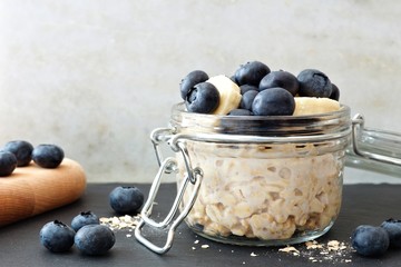 Jar of overnight oats with fresh blueberries and bananas, scene with white and black stone...