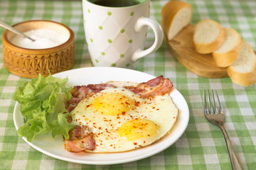 Fried two eggs with bacon and lettuce, white bread, tea and sugar on a tablecloth in a green cage.