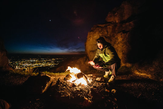 young male hiker crouching at a campfire at night on a mountain