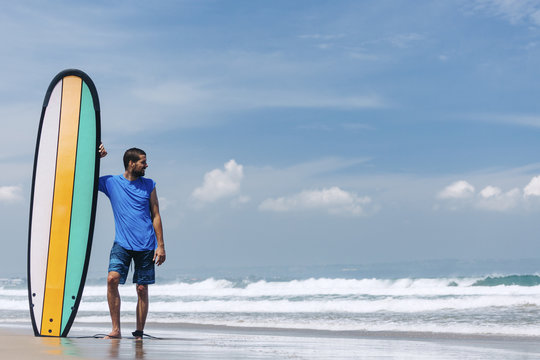 Portrait of a surfer standing next to longboard on the beach, looking at the surfing waves