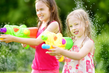 Adorable little girls playing with water guns on hot summer day. Cute children having fun with water outdoors.
