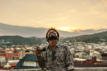 Young black man laughing holding an old camera on a rooftop at sunset.