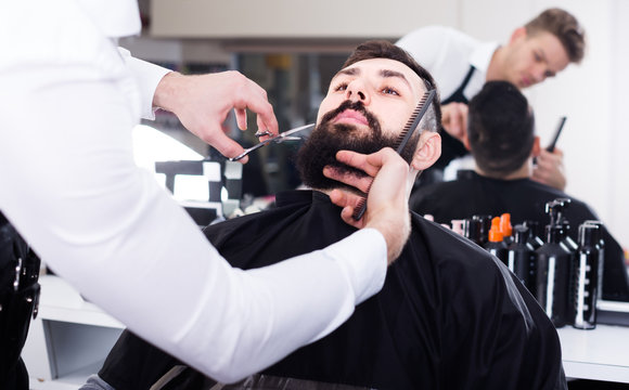 Cheerful man forming beard of client into shape