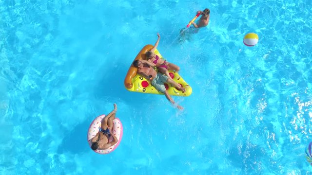 AERIAL: Cheerful friends having water fight on colorful floaties at pool party. Playful guys and girls splashing water and taking selfies on inflatable pizza, doughnut, flamingo and watermelon floats