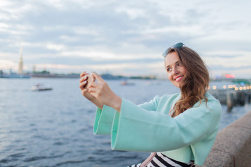 Fototapeta na wymiar Young and beautiful girl sitting on the embankment of the river. she looks at the sunset and taking a selfie on your phone. Saint Petersburg, Russia