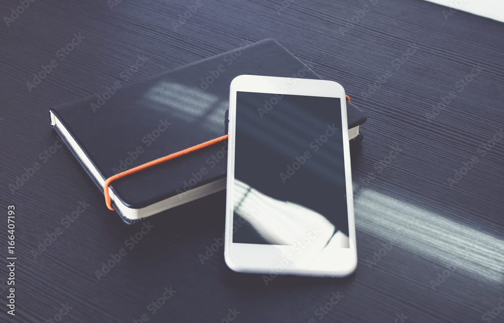 Aufkleber blank screen of mobile phone lay on black notebook near window on black wooden modern desk at aftern