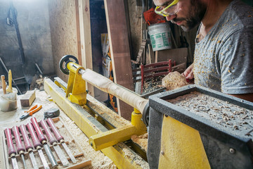 A dark-haired man with a beard and protective green glasses makes a wooden product on a lathe; work on a lathe; a lot of wooden sawdust fly apart