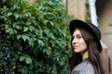 Headshot of fashionable attractive brunette girl with cute smile wearing stylish black hat posing outdoors, standing at green wall, spending weekend in city park. People, leisure and lifestyle concept