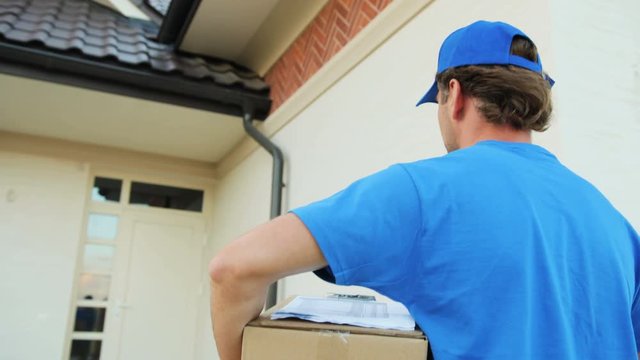 Close up portrait of courier walking with cardboard to the house for delivering package to the owner. View from the back.