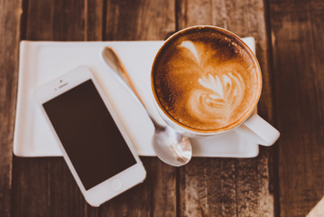 Cup of latte coffee with white modern smartphone on old rustic wooden table. Top view