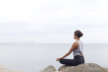Woman seated in a lotus yoga pose on rocks beside a bay