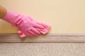Hand in rubber protective glove with microfiber cloth cleaning baseboard on the floor from dust at...