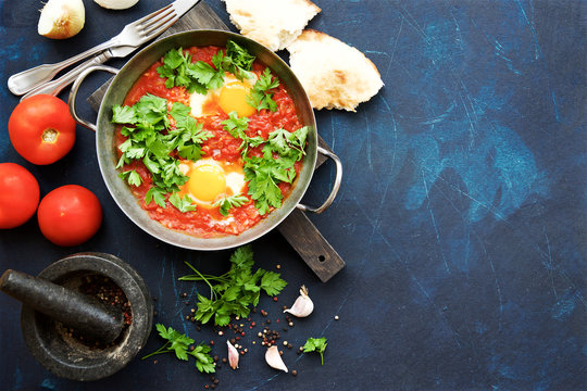 Shakshuka fried eggs with tomatoes, onions and spices in frying pan on a dark blue cracked background  
