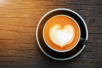 Heart shape latte art coffee in black cup on old wooden desk warm light and flare effect , top view and copy space