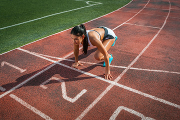 Young woman runner with dark hair in a white sports top and short shorts stands in a low start...