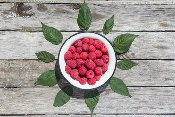 Raspberry in a white plate on an aged wood background. Green raspberry leaves. Top view.