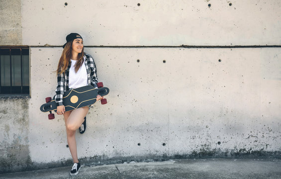 Portrait of happy young woman with longboard standing in front of concrete wall