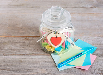 Glass jar with handmade wooden hearts decorations and ribbon near a stack of colored papers and a blue pen.