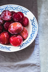 Plums of different varieties in a white ceramic plate with a pattern. Gray concrete background, gray textile napkin.Top view. Agriculture, Gardening, Harvest Concept.