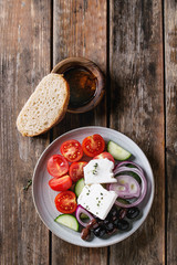 Ingredients for traditional greek salad. Cherry tomatoes, sliced cucumbers, red onion, black olives, feta cheese gray plate with loaf bread, olive oil over wooden plank background. Top view