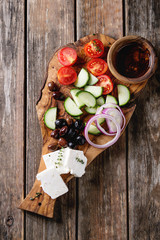 Ingredients for traditional greek salad. Cherry tomatoes, sliced cucumbers, red onion, black olives, feta cheese on olive wood board with olive oil over wooden plank background. Top view