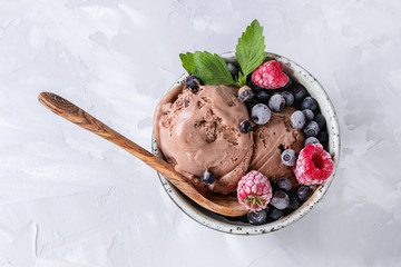 Homemade chocolate ice cream with frozen berries blueberry, raspberry, mint served in white bowl with olive wood spoon over gray concrete background. Top view with space