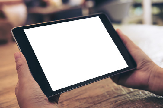 Mockup image of hands holding black tablet pc with white blank screen on wooden table background in cafe