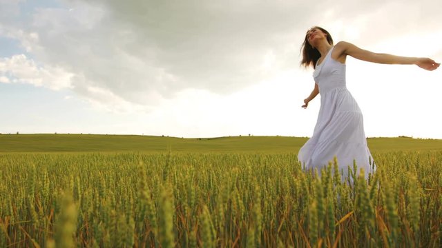 Elegant young woman in the long white dress dancing in yellow wheat field on sky background.