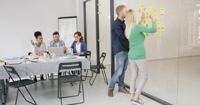 Group of people in modern office and two of them brainstorming on new plan using stickers and building diagram on glass wall.