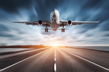 Airplane and road with motion blur effect at sunset. Landscape with passenger airplane is flying over the asphalt road and cloudy sky. Commercial plane is landing. Aircraft with blurred background  - obrazy, fototapety, plakaty